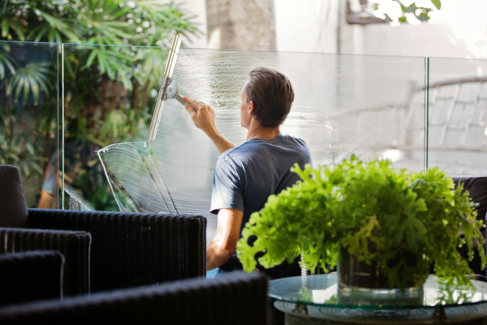 A man cleaning a glass barrier outdoors with a squeegee, surrounded by greenery.